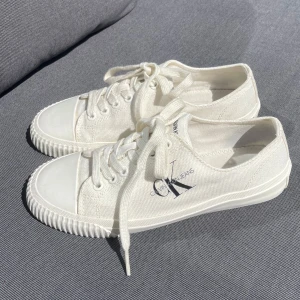 Vita sneakers från Calvin Klein Jeans - Snygga vita sneakers från Calvin Klein Jeans med klassisk låg profil och rund tå. Skorna har snörning, platt sula och är tillverkade i canvas med logga på sidan och hälen. Perfekta för en clean och stilren look.Vita sneakers från Calvin Klein Jeans. Använda 1 gång så i bra skick med visst slitage på undersidan av sulan samt minimala missfärgningar på tyget, var god se bilder. Storlek 37, normala i storleken.