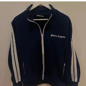 Blå tröja Palm Angels - Snygg marinblå tröja från Palm Angels med vita ränder längs ärmarna och dragkedja framtill. Jackan har hög krage och logga på bröstet. Perfekt för dig som gillar streetwear och vill sticka ut med en sportig look.