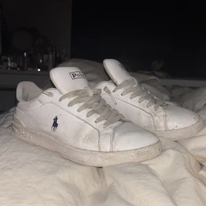 Vita sneakers från Polo Ralph Lauren - Klassiska vita sneakers från Polo Ralph Lauren med låg profil och rund tå. Skorna har snörning, diskret Polo-logga på sidan och märkesdetaljer på plös och sula. Tillverkade i skinn med platt sula, perfekta för en clean och stilren look.