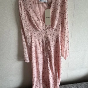 Ljusrosa blommig klänning från Adoore - Supersöt ljusrosa långklänning från Adoore med små blommiga mönster, v-ringning och knappar framtill. Klänningen har långa ärmar och en feminin passform som ger en romantisk vibe. Perfekt för vår och sommar!