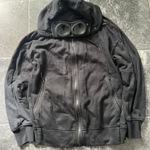 Svart hoodiejacka från C.P. Company - Svart hoodiejacka från C.P. Company med ikoniska goggles i huvan. Jackan har dragkedja framtill, två fickor med dragkedja och ribbade muddar. Tillverkad i mjuk bomull, perfekt för en streetwear-look. Priset kan diskuteras vid snabb affär 