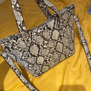 Axelväska i ormmönstrat skinn Dondonna - Snygg axelväska från Dondonna i ormmönstrat skinn. Väskan har både handtag och justerbar axelrem, dragkedja upptill och flera fack inuti. Färgerna går i beige, svart och grått vilket ger en cool och trendig look.