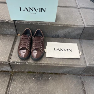 Bruna sneakers från Lanvin i mocka - Passar 41-43Påse box osv följer med Snygga bruna sneakers från Lanvin med ovandel i mocka och lackad tå. Skorna har svarta platta sulor och klassisk snörning. Perfekta för dig som gillar exklusiva och stilrena sneakers med en lyxig touch. 