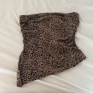 Leopardmönstrad tubtopp i brunt - Säljer en tubtopp med leopardmönster i bruna och svarta toner. Toppen har en asymmetrisk nederkant som ger en cool twist och är axelbandslös för en trendig look. 