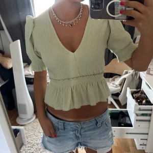Ljusgrön blus med volang och v-ringning - Supersöt ljusgrön blus med v-ringning, puffiga korta ärmar och volang nedtill. Toppen har en smockad midja som ger en snygg siluett och är gjord i ett lätt, krinklat tyg. Perfekt för dig som vill ha en feminin och trendig look.