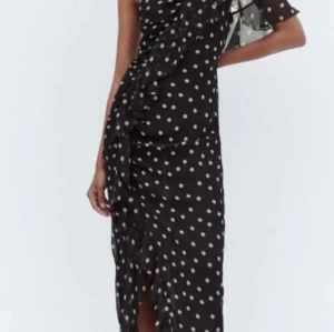Svart prickig långklänning från Zara - Supersnygg svart långklänning från Zara med vita/ljusrosa prickar och volangdetaljer längs framsidan. Klänningen har oneshoulder-design med en kort ärm och en axelbandslös sida. Materialet är lätt och luftigt, perfekt för att sticka ut med en cool look.