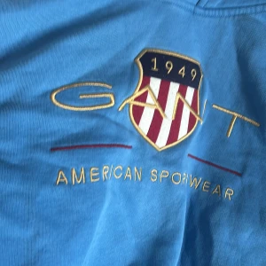 Blå hoodie från GANT med brodyr - Snygg blå hoodie från GANT med broderad logga och amerikanskt sköldmärke på bröstet. Tröjan har huva med snörning, känguruficka och ribbade muddar. Perfekt för dig som gillar sportig och avslappnad stil.