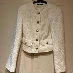 Vit kavajjacka från Gina Tricot - Stilren vit kavajjacka från Gina Tricot med guldknappar framtill och på fickorna. Jackan är kortare i modellen, har rund halsringning och är tillverkad i ett strukturerat tyg. Storlek 34 i nyskick då den knappt är använd och inga fläckar.