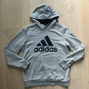Grå hoodie från Adidas med logga - Säljer en grå hoodie från Adidas med stor svart logga på bröstet. Tröjan har huva, magficka och ribbade muddar vid ärmslut och nederkant.