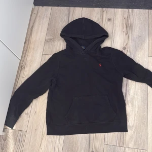Svart hoodie från Polo Ralph Lauren - Svart hoodie från Polo Ralph Lauren med klassisk röd broderad logga på bröstet. Tröjan har huva, känguruficka och är tillverkad i mjuk bomull. Perfekt för chill dagar och enkel att matcha med jeans eller sweatpants. Priset ej fast, hör av er vid minsta lilla!