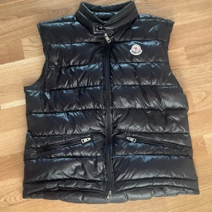 Äkta Moncler Gui Väst | 1-S - Snygg svart dunväst från Moncler med glansig finish och klassisk logga på bröstet. Västen har ståkrage, två dragkedjeförsedda fickor och quiltad design. Perfekt att slänga över en hoodie för extra värme. Stängs med dragkedja framtill. Inga defekter jätte snygg, NFC Scan och QR kod funkar, Nypris 9290kr mitt pris 3450kr, passar 165-177