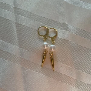 Pearly spike - Snygga örhängen i guld med rund vit pärldetalj och spetsiga hängen. De har en cool och modern design som sticker ut, perfekta för att lyfta din stil. Lätta att bära med smidig låsning och trendig känsla.