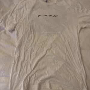 Vit t-shirt med Manhattan-tryck - Vit t-shirt i bomull med svart text 'From The Rooftops' på bröstet och ett stort tryck med Manhattan skyline på ryggen. Snyggt street-inspirerat motiv och klassisk passform med korta ärmar. Perfekt för dig som gillar urban stil och coola prints.