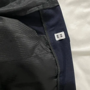 Mörkblå kostymbyxor Jack & Jones - Snygga mörkblå kostymbyxor från Jack & Jones med klassisk passform och diskret metallogo vid fickan. Byxorna har hällor för bälte och är tillverkade i en mjuk bomullsblandning som känns skön hela dagen. Perfekta för en clean och stilren look.