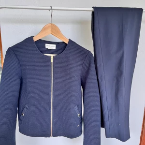 Mörkblått set från Holly & White - Snyggt mörkblått set från Holly & White med ribbad tröja och matchande byxor. Tröjan har rund hals, guldfärgad dragkedja och fickor med metallknappar. Byxorna har klassisk passform och diskret prickigt foder i midjan. Perfekt för dig som gillar stilrena detaljer. Tröjan är S. Byxorna är 38.