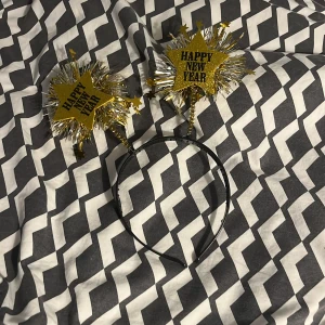 Glittrigt nyårsdiadem med stjärnor - Säljer ett svart diadem med två stora glittriga stjärnor i guld och silver där det står 'Happy New Year'. Stjärnorna är dekorerade med glittriga paljetter och små stjärnor som sticker ut, perfekt för nyårsfirandet. Diademet är gjort av syntetmaterial och har en festlig look.