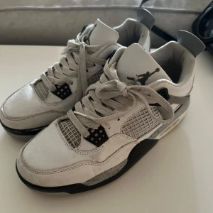 Nike Air Jordan 4 vit/grå sneakers - Nike Air Jordan 4 sneakers i vit och grå färg med svarta detaljer. Skorna har klassisk snörning, meshpaneler och en synlig Air-sula. Ovandelen är i skinn med texturerad yta och coola plastdetaljer på sidorna. Perfekt för dig som gillar streetstyle och ikoniska sneakers. De är inte äkta!!!! 