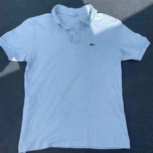 Ljusblå Lacoste piké t-shirt - Fräsch ljusblå piké från Lacoste med klassisk krage och två knappar vid halsen. Ikoniska krokodilloggan broderad på bröstet. T-shirten är i bomull och har korta ärmar, perfekt för dig som gillar stilren och sportig vibe.