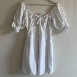 Vit kort klänning från NLY Trend - Supersöt vit kortklänning från NLY Trend med puffärmar och knyt framtill vid halsringningen. Klänningen har en feminin passform och är gjord i mjuk bomull, perfekt för varma dagar. Den har en dragkedja i sidan för enkel påklädning och en snygg, lite romantisk stil.