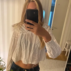 Vit blus med volang från Gina Tricot - Superfin vit blus från Gina Tricot med volangdetaljer på axlarna och spetsinslag. Lätt och luftig bomull, v-ringad och med långa ärmar med resår vid ärmslut. Den har tyvär blivit för kort för mig och det är därför jag säljer den💕😊