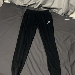 Svarta mjukisbyxor från Nike - Svarta mjukisbyxor från Nike med vit logga på vänster lår. Byxorna har dragsko i midjan och muddar vid bensluten för en skön och avslappnad passform. Perfekta för chill eller träning.