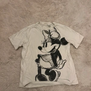 Vit t-shirt med Minnie Mouse-tryck - Vit t-shirt från Disney med ett stort, skissat tryck av Minnie Mouse på framsidan. Klassisk rund hals och korta ärmar. Mjuk bomullskvalitet som är skön mot huden. Perfekt för dig som gillar coola och lekfulla motiv.