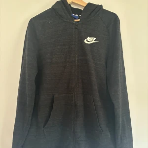 Svart hoodiejacka från Nike, stl S - Svart hoodiejacka från Nike med dragkedja framtill och klassisk logga på bröstet. Jackan har huva, två fickor och är tillverkad i ett mjukt, strukturerat material. Perfekt för en sportig och avslappnad stil.