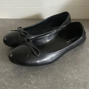 Svarta ballerinaskor med rosett - Klassiska svarta ballerinaskor från Åhléns i skinnimitation. Skorna har rund tå och en söt liten rosett framtill. Perfekta för en stilren och enkel look, passar till både jeans och klänning.