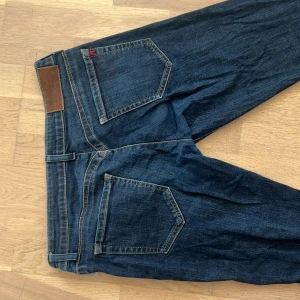 Replay mörkblå jeans straight fit - Mörkblå jeans från Replay med raka ben och klassisk femficksdesign. Jeansen har guldiga detaljer vid knappen, kontrastsömmar och Replay-logga på fickan. Tillverkade i slitstarkt denim för en tidlös look.