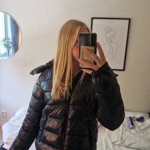 Svart pufferjacka med huva från 157 - Säljer en svart pufferjacka från 157 med glansig finish och huva. Jackan har dragkedja framtill, två fickor med dragkedja och quiltad design. Perfekt för kalla dagar och riktigt skön att bära. Passformen är normal och jackan är långärmad. Jättefin och snygg till höst, vinter och vår💕 trendig❤️