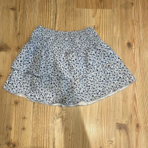 Blommig volangkjol från Gina Tricot - Supersöt kort kjol från Gina Tricot med blått blommönster på vit botten. Kjolen har volanger och en smockad midja som gör den bekväm och lätt att bära.💞💞