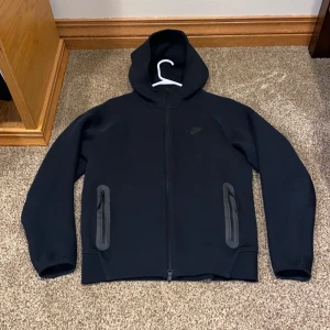 Svart Nike Tech Fleece hoodie XS - Nike Tech Fleece hoodie i svart med hel dragkedja och två fickor med dragkedja. Snygg minimalistisk design med diskret Nike-logga på bröstet. Jackan har huva och är tillverkad i mjuk fleece för extra komfort. Perfekt för chill eller när du är på språng. Dragkedjan funkar inte att dra isär längst nere! Därav säljer jag billigt!