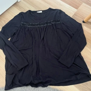 Svart långärmad blus från Pompdelux - Svart långärmad blus från Pompdelux med söt volangdetalj framtill och rund halsringning. Mjuk och skön i bomull, perfekt för dig som gillar en enkel men ändå stylish look. Passar till både jeans och kjol.