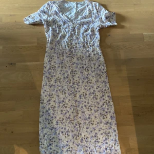 Ljus blommig långklänning med knappar - Säljer en romantisk långklänning med ett slit på botten och lila, beige och bruna blommor. Klänningen har korta ärmar, v-ringning och knappar framtill. Materialet känns som mjuk bomull, perfekt för varma dagar. Använd 1 gång. Pris går att förhandla.