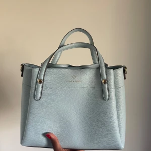 Ljusblå handväska från Nanette Lepore - Snygg ljusblå handväska från Nanette Lepore i stilren design. Väskan har guldiga metalldetaljer och är gjord i strukturerat ”skinn”. Perfekt storlek för att få plats med allt du behöver och passar till många olika outfits. Finns även ett längre band till ,om man vill bära den på andra sätt💕