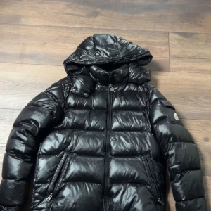 Svart Moncler dunjacka med huva - Snygg svart dunjacka från Moncler med glansig finish och huva. Jackan har dragkedja framtill, två fickor med dragkedja och klassisk Moncler-logga på ärmen. Perfekt för kalla dagar när du vill hålla stilen och värmen.