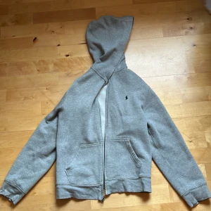 Grå hoodie från Polo Ralph Lauren storlek 158-164 - Klassisk grå hoodie från Polo Ralph Lauren med dragkedja framtill, huva och två stora fickor. Tröjan har en liten broderad logga på bröstet och är tillverkad i mjuk bomullsmix. Perfekt för en chill och avslappnad stil.