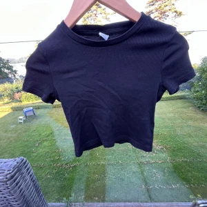 Svart ribbad topp från Lindex - En svart kortarmad tröja  från Lindex med ribbad struktur och rund hals. Modellen är kortärmad och har en slim passform som sitter snyggt. Perfekt för dig som gillar enkel och stilren look. Materialet är mjuk bomull som känns skönt mot huden.