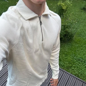 Vit half zip tröja från East West - Säljer en stilren vit långärmad tröja från East West i storlek M. Tröjan har en snygg krage och en half zip-dragkedja framtill. Materialet är mjuk bomull med lätt ribbad struktur, perfekt för dig som gillar enkel och clean look.
