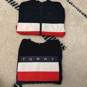 Tommy Hilfiger tröja med logga - Snygg svart tröja från Tommy Hilfiger med stor logga och breda blockränder i vitt och rött framtill. Klassisk rund hals och ribbade muddar. Perfekt för dig som gillar streetstyle och vill sticka ut med en ikonisk look.