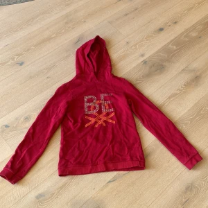 Röd hoodie från United Colors of Benetton - Snygg röd hoodie från United Colors of Benetton med glittrande BE-text och dekorativa nitar på bröstet. Tröjan har huva, långa ärmar och är gjord i mjuk bomull. Perfekt för dig som vill ha en cool och bekväm look.