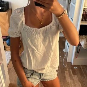 Vit puffärmad blus med fyrkantig ringning - Supersöt vit blus med puffärmar och fyrkantig ringning. Toppen har resår vid ärmslut och hals, vilket ger en romantisk vibe. Perfekt till shorts eller kjol för en fräsch look. Luftigt material som passar när det är varmt ute.