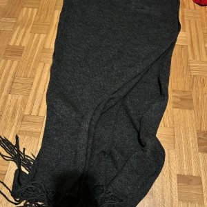 Mörkgrå stickad halsduk med fransar - En riktigt mysig och lång mörkgrå halsduk med fransar i ändarna. Perfekt att vira flera varv runt halsen för extra värme. Stilren och enkel design som passar till det mesta. Materialet är stickat och känns mjukt mot huden.