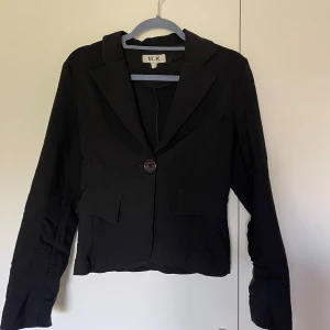 Svart kavaj från M.K med knapp - Stilren svart kavaj från M.K med klassisk krage och en knapp framtill. Modellen är kortare och har två fejk fickor med lock. Perfekt för dig som gillar en enkel och snygg look. Passar till många olika outfits och har en tidlös design.