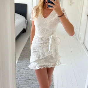 Vit spetsklänning   - Supersöt vit kortklänning från Dennis Maglic med blommigt spetsmönster. Klänningen har omlottkänsla med knyt i midjan, volangdetaljer och dragkedja bak. Perfekt som konfirmationsklänning eller studentklänning! (Aldrig använd!)