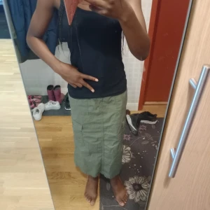 Grön cargo långkjol med fickor - Säljer en grön långkjol i cargo-stil med stora fickor på sidorna och knappdetaljer. Kjolen har en rak passform och är gjord i bomull, perfekt för dig som gillar utility-vibe och streetstyle. Midjan har hällor för bälte och kjolen har slits bak för extra rörelsefrihet. (För åldrarna 12-13 år)