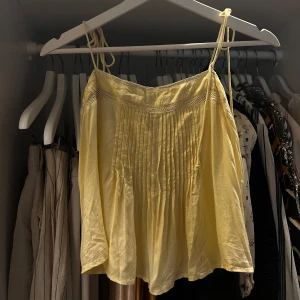 Gul linnetopp från Urban Outfitters - Supersöt ljusgul linnetopp från Urban Outfitters med tunna axelband man kan knyta/juster själv och fina spetsdetaljer upptill. Toppen har plisserad framsida och är luftig med en flowy passform, perfekt för varma dagar. Tillverkad i bomull och har en romantisk vintagekänsla.
