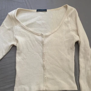 Ribbad beige topp från Brandy Melville - Superfin ribbad topp från Brandy Melville i beige med lång ärm och knappar hela vägen framtill. Den har en croppad passform och djup rundad ringning. Perfekt att styla med jeans eller kjol för en chill look. Säljer en likadan i svart 💕