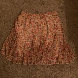 Blommig kortkjol från Amisu - Supersöt kortkjol från Amisu i storlek 34 med blommigt mönster i rött, gult och grönt på vit botten. Kjolen har volangkant och är gjord i ett lätt och luftigt material, perfekt för varma dagar. Elastisk midja för skön passform.