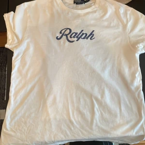 Vit t-shirt från Polo Ralph Lauren - Vit t-shirt från Polo Ralph Lauren med klassisk passform och blått 'Ralph'-tryck på bröstet. T-shirten har rund hals och korta ärmar, tillverkad i mjuk bomull som känns skön mot huden. Perfekt för en chill och stilren look.